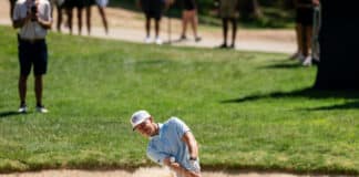 Golfer wearing a light blue shirt and white cap explodes from a sand bunker while spectators watch from the grassy edge (blurry background).