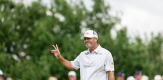 Male golfer in a white polo and cap waves to cheering spectators during a tournament.
