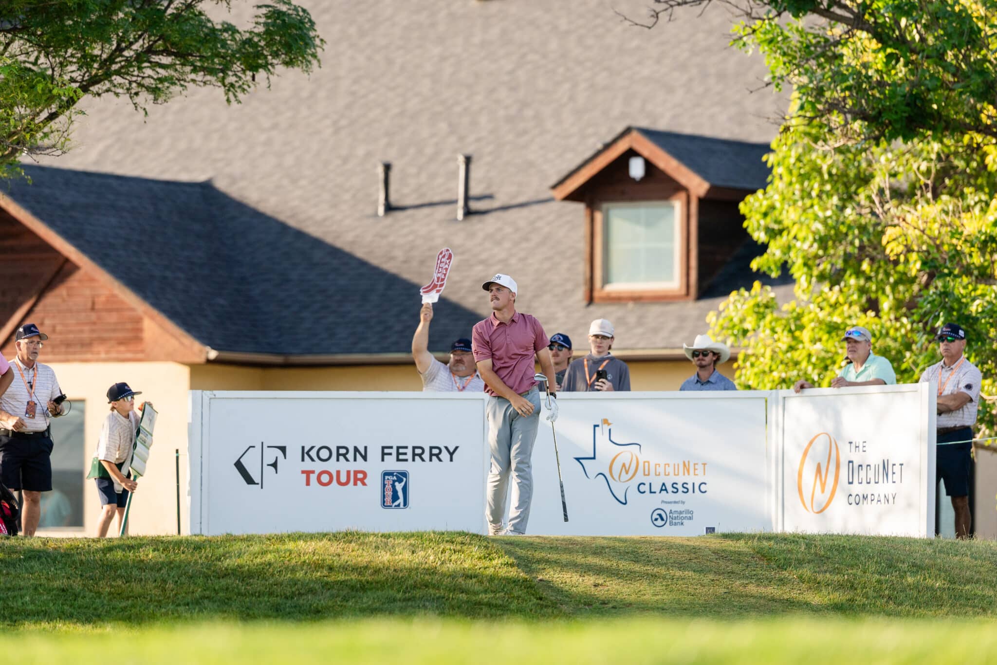 Golfer in a pink shirt raises his club on a raised platform during a tour event, with banners reading Korn Ferry Tour and OccuNet Classic and spectators in the background.