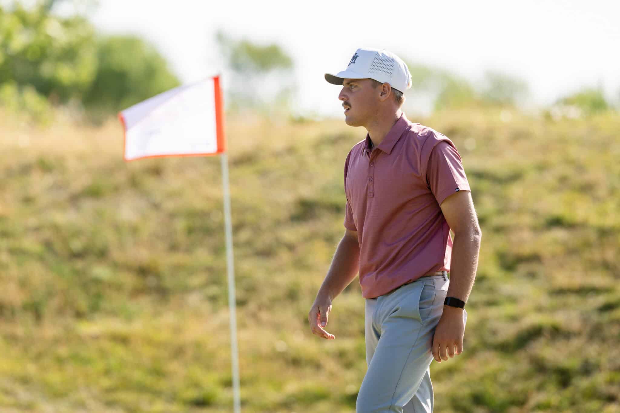 Man wearing a white cap and pink polo walks along a sunny course near a red-bordered flag marker.