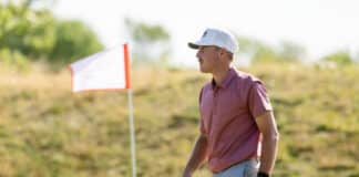 Man wearing a white cap and pink polo walks along a sunny course near a red-bordered flag marker.