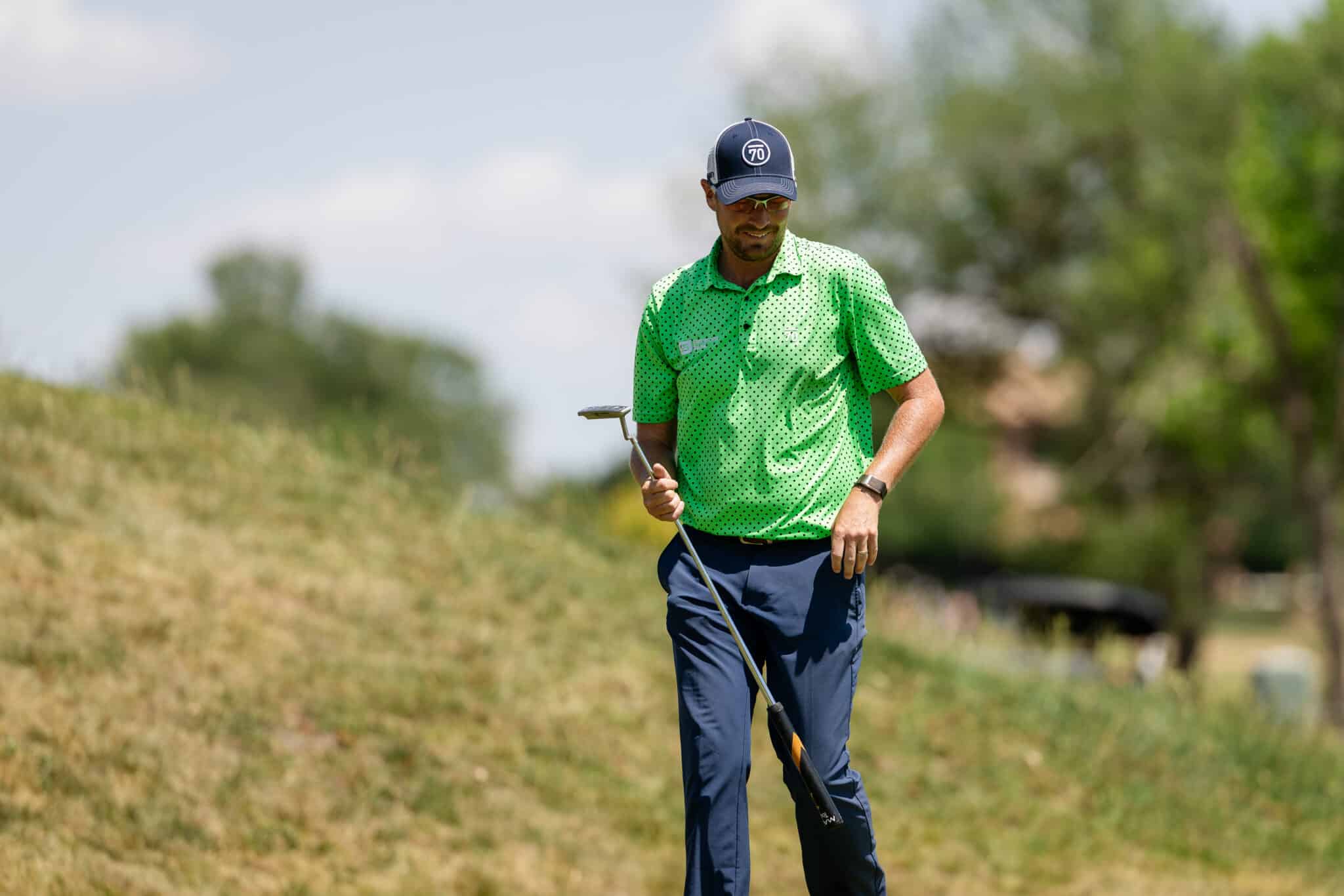 Male golfer on a sunny course wearing a green polka-dot polo and navy cap, holding a club ready to swing.