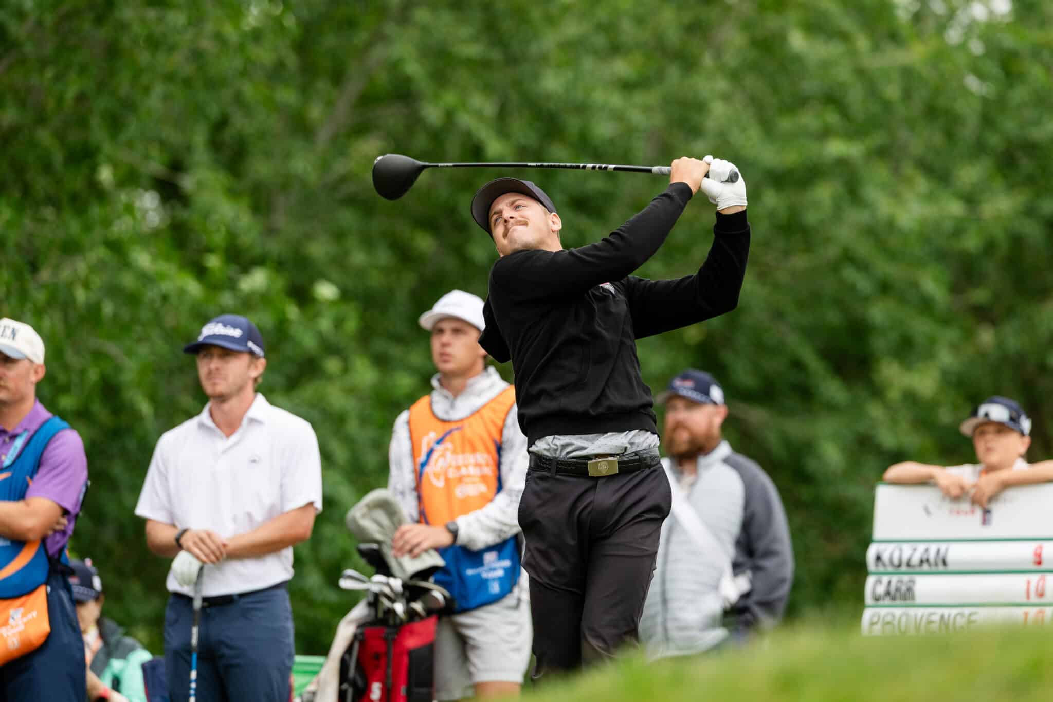 korn-ferry-tour-occunet-classic-day-5-jpegJoe-213 | Press Pass Sports Man in black clothes swinging a golf club mid-shot, with spectators and greenery in the background.