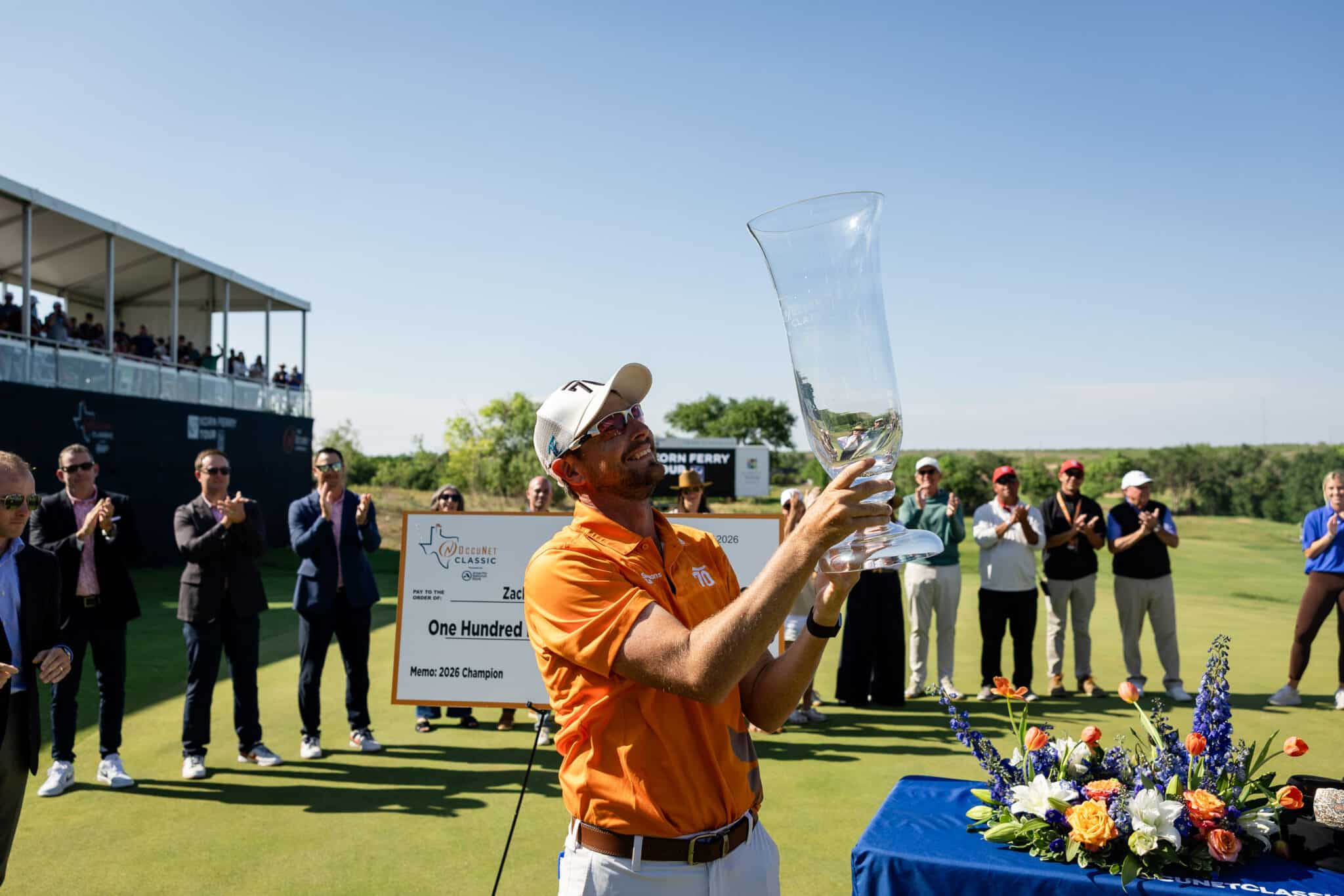korn-ferry-tour-occunet-classic-day-5-jpegJoe-592 | Press Pass Sports Man in an orange shirt holds a tall crystal trophy aloft as spectators applaud at an outdoor award ceremony, with flowers in the foreground.