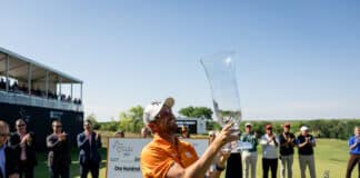 Man in an orange shirt holds a tall crystal trophy aloft as spectators applaud at an outdoor award ceremony, with flowers in the foreground.