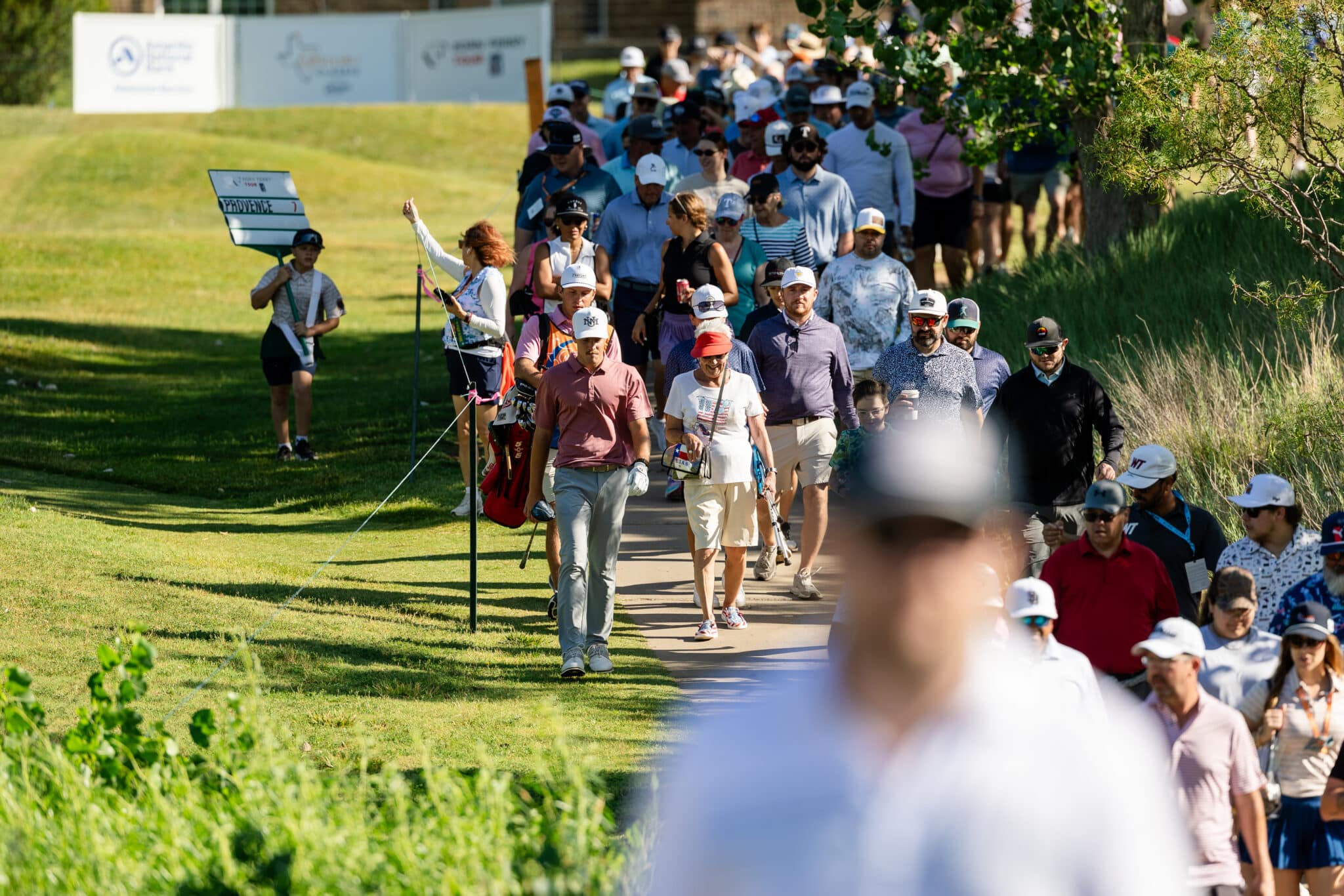 korn-ferry-tour-occunet-classic-day-3-jpegJoe-80 | Press Pass Sports A crowd of golfers walking along a sunny golf course path, many wearing hats and carrying clubs with volunteers nearby.
