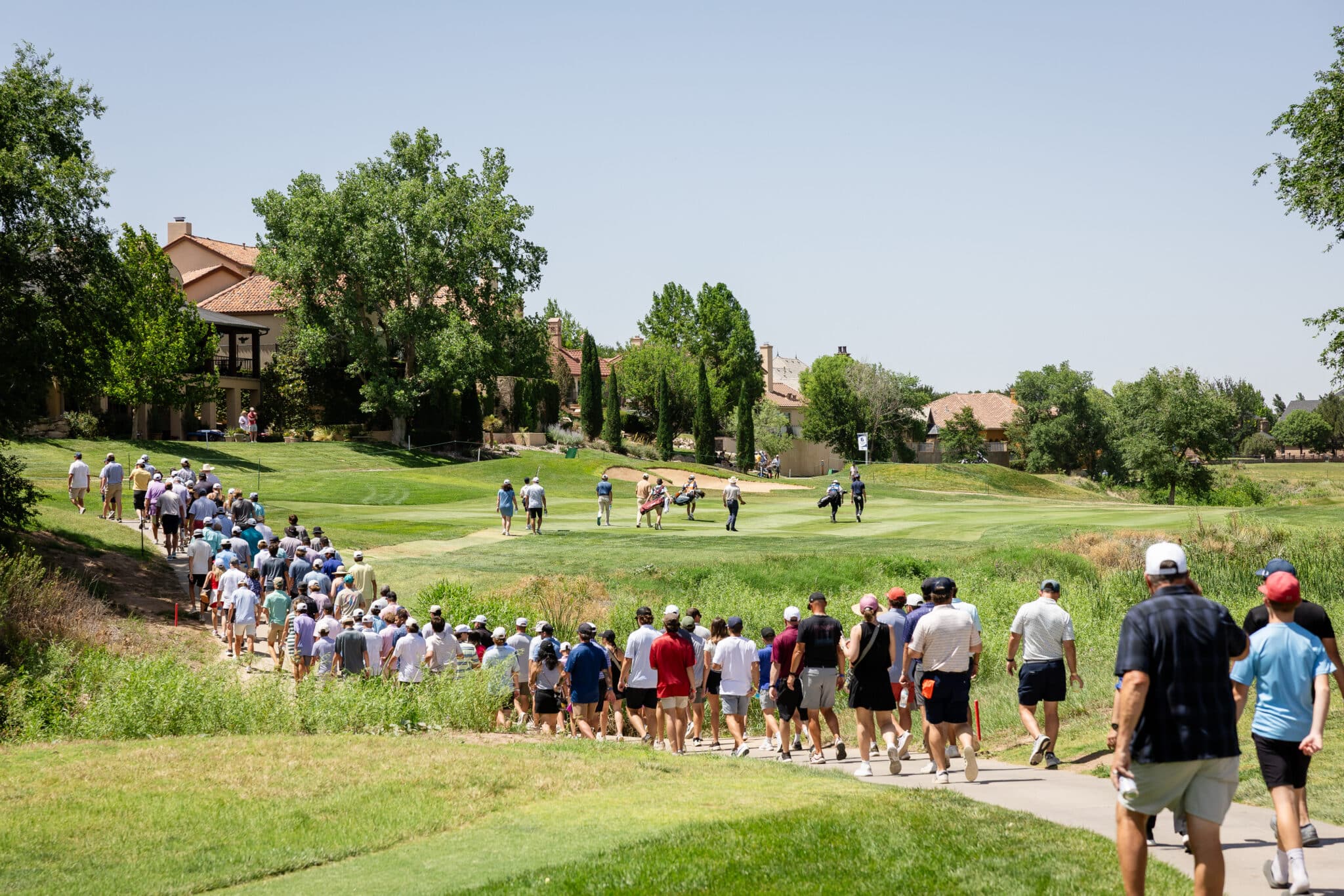 korn-ferry-occunet-classic-day-1-jpegJoe-310 | Press Pass Sports A large group of people walking along a paved path across a sunny golf course, with green grass and houses in the background.