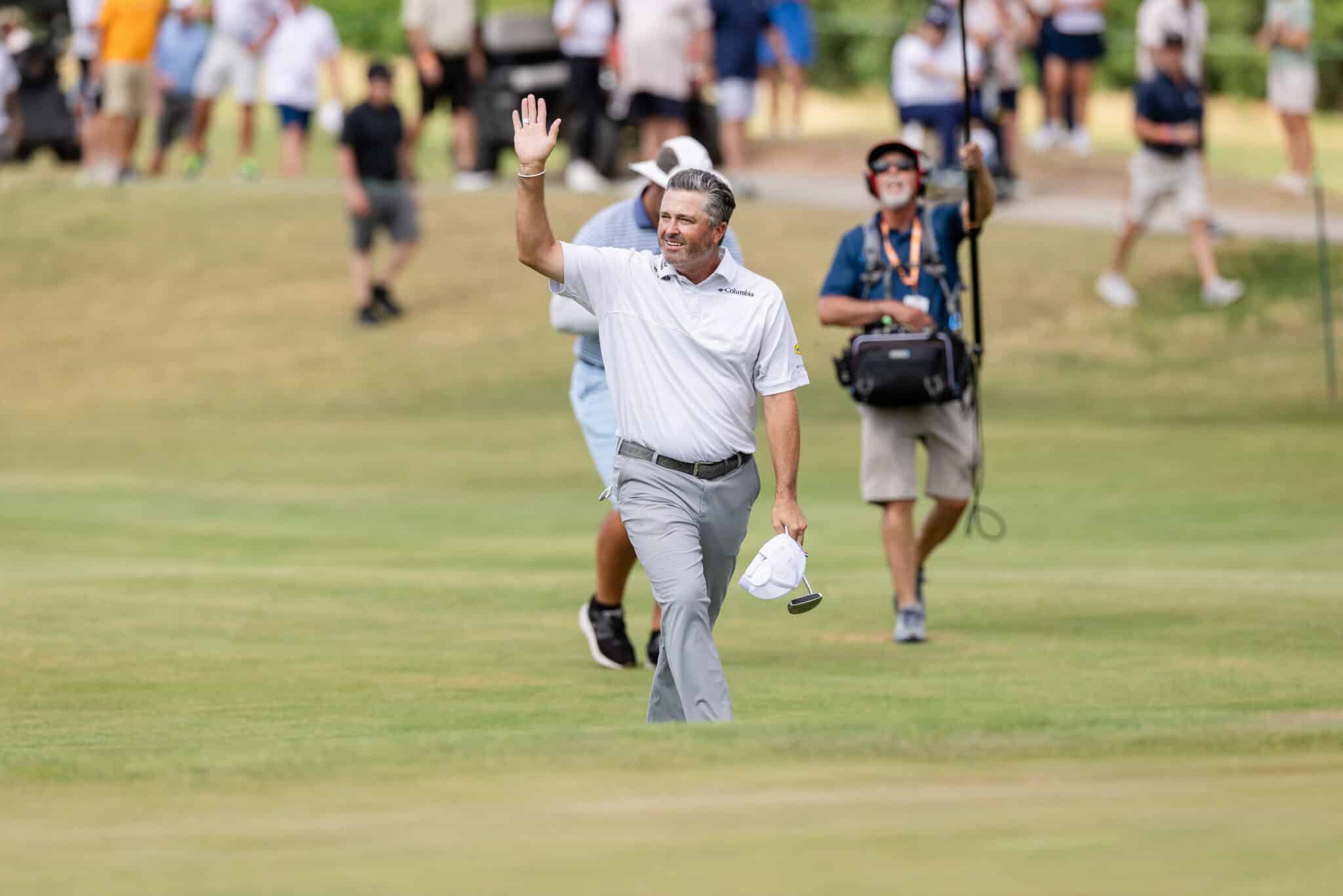 korn-ferry-occunet-classic-day-2-jpegJoe-478 | Press Pass Sports Man in a white polo waves to the crowd while walking on a golf course, holding a hat; blurred spectators in the background.