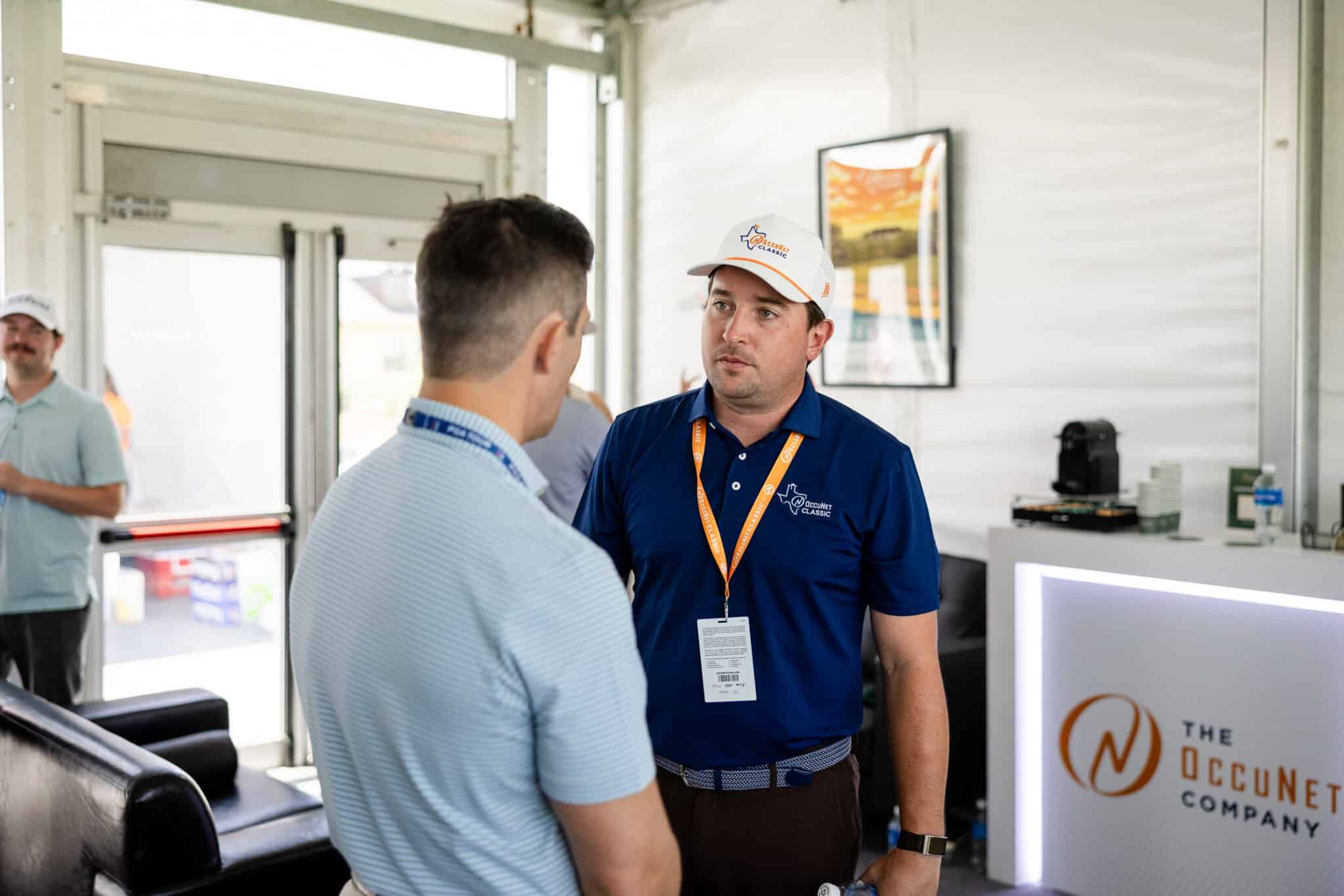 korn-ferry-occunet-classic-day-1-jpegJoe-237 | Press Pass Sports Two men converse at a trade show booth; one wearing a white cap and blue polo with an orange lanyard stands near a branded display panel on the right.
