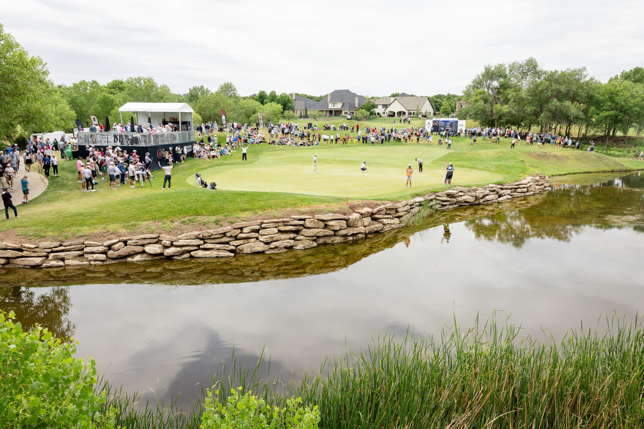 korn-ferry-tour-occunet-classic-day-5-jpegJoe-299 | Press Pass Sports Crowd gathered around a golf green near a stage with a white canopy, overlooking a stone-lined water hazard and a sunny suburban backdrop.