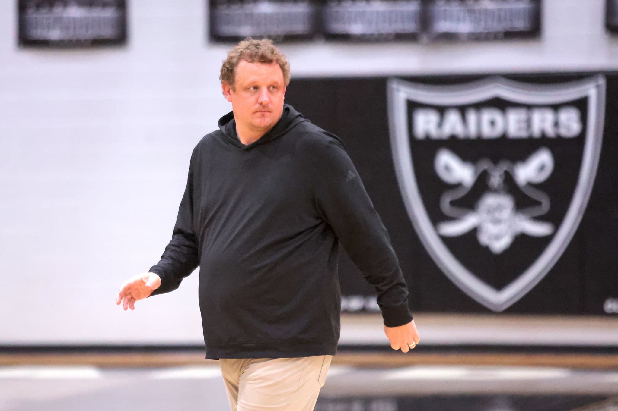 Man in a black hoodie walking indoors with a Raiders banner in the background.