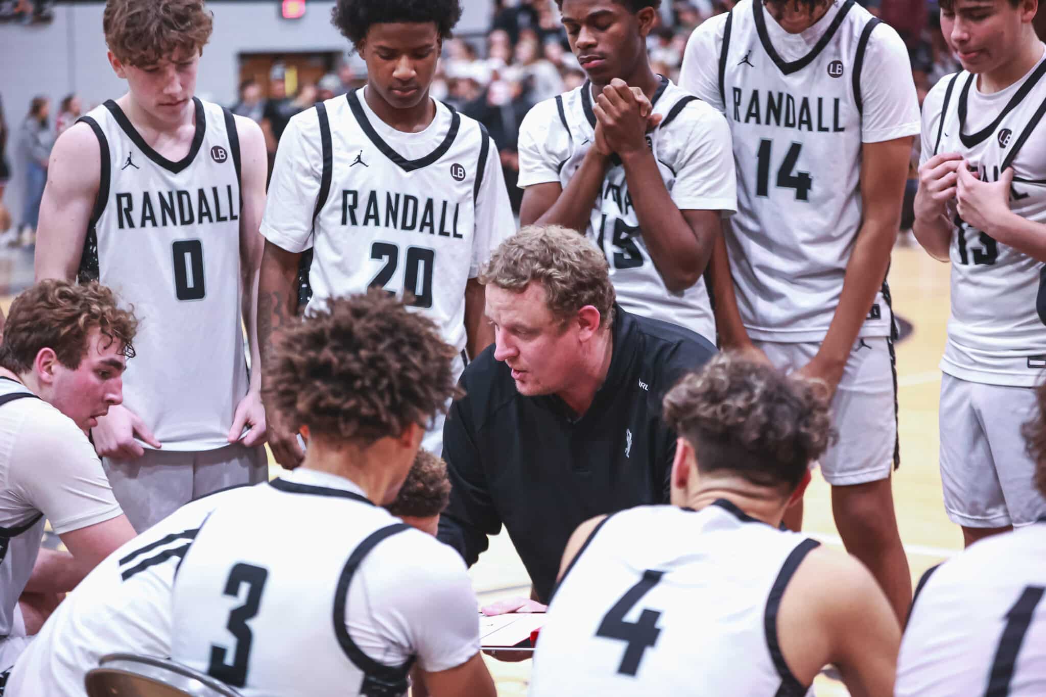MY4A9876 | Press Pass Sports Basketball team in white RANDALL uniforms gathers around coach for a huddle on the court during a timeout.