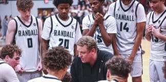 Basketball team in white RANDALL uniforms gathers around coach for a huddle on the court during a timeout.