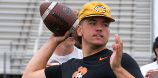 Young man in a black T-shirt with an orange graphic throws a football behind his head outdoors, wearing a yellow cap.