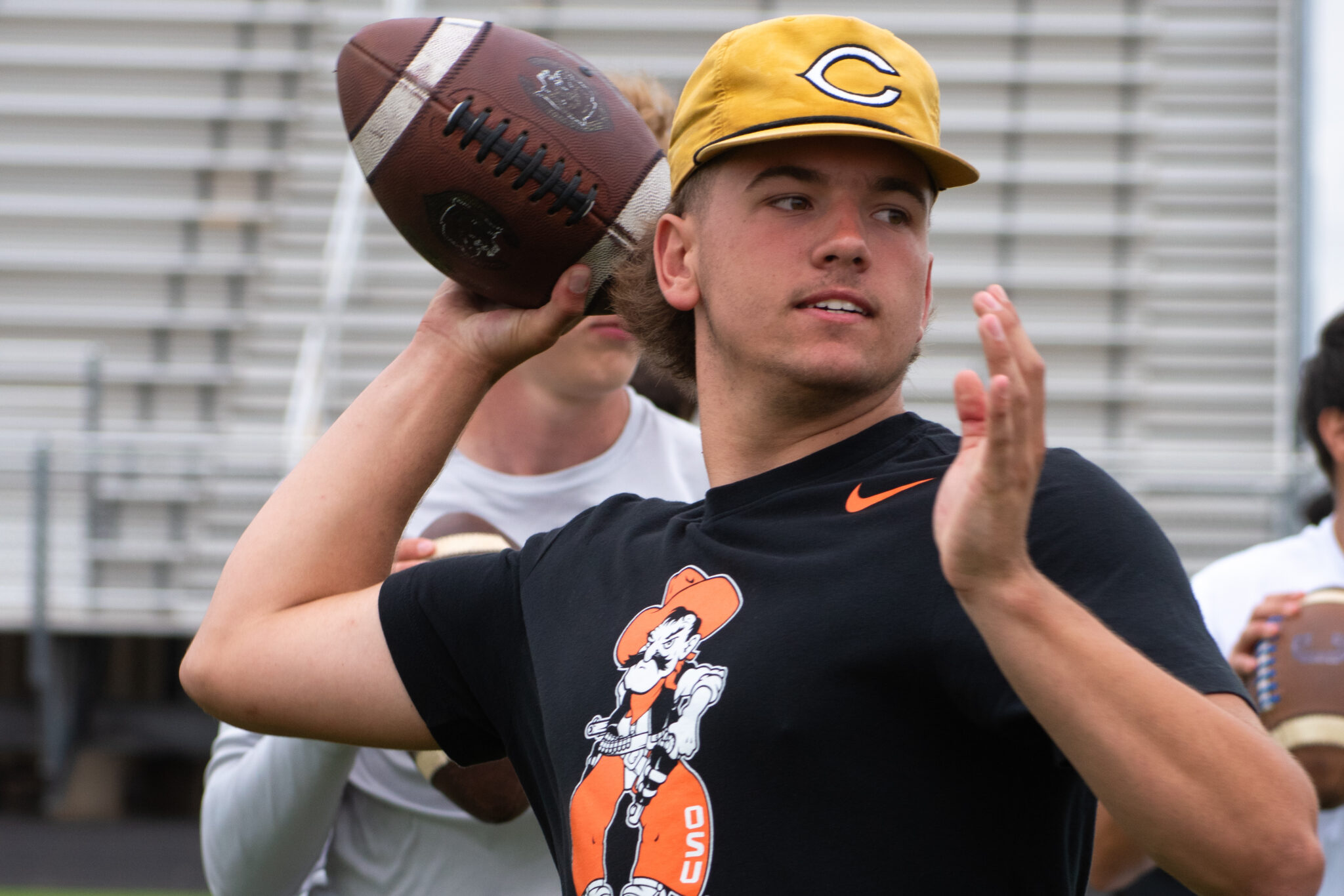 DSC_0019 | Press Pass Sports Young man in a black T-shirt with an orange graphic throws a football behind his head outdoors, wearing a yellow cap.