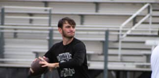 Young man in a black shirt throwing a football on a sports field with empty bleachers in the background]