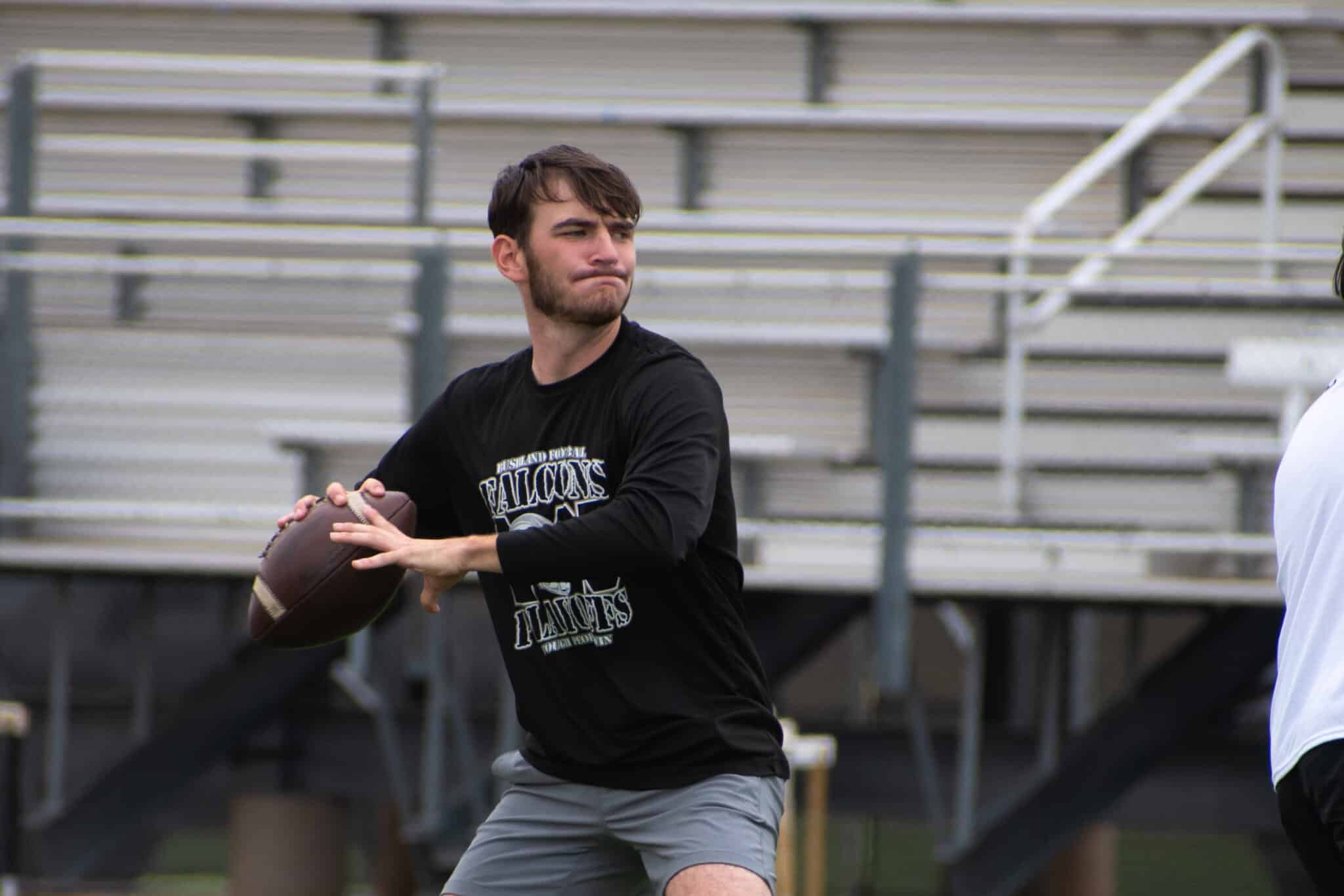 Young man in a black shirt throwing a football on a sports field with empty bleachers in the background]