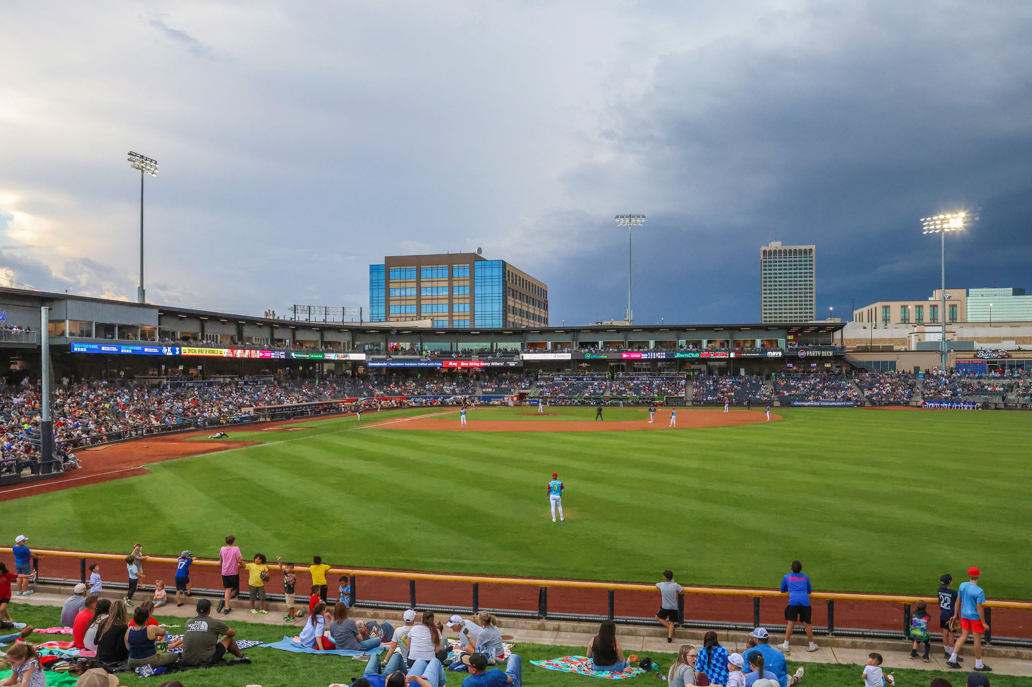 Crowd watching a baseball game at a stadium with tall floodlights and cloudy sky in the background.