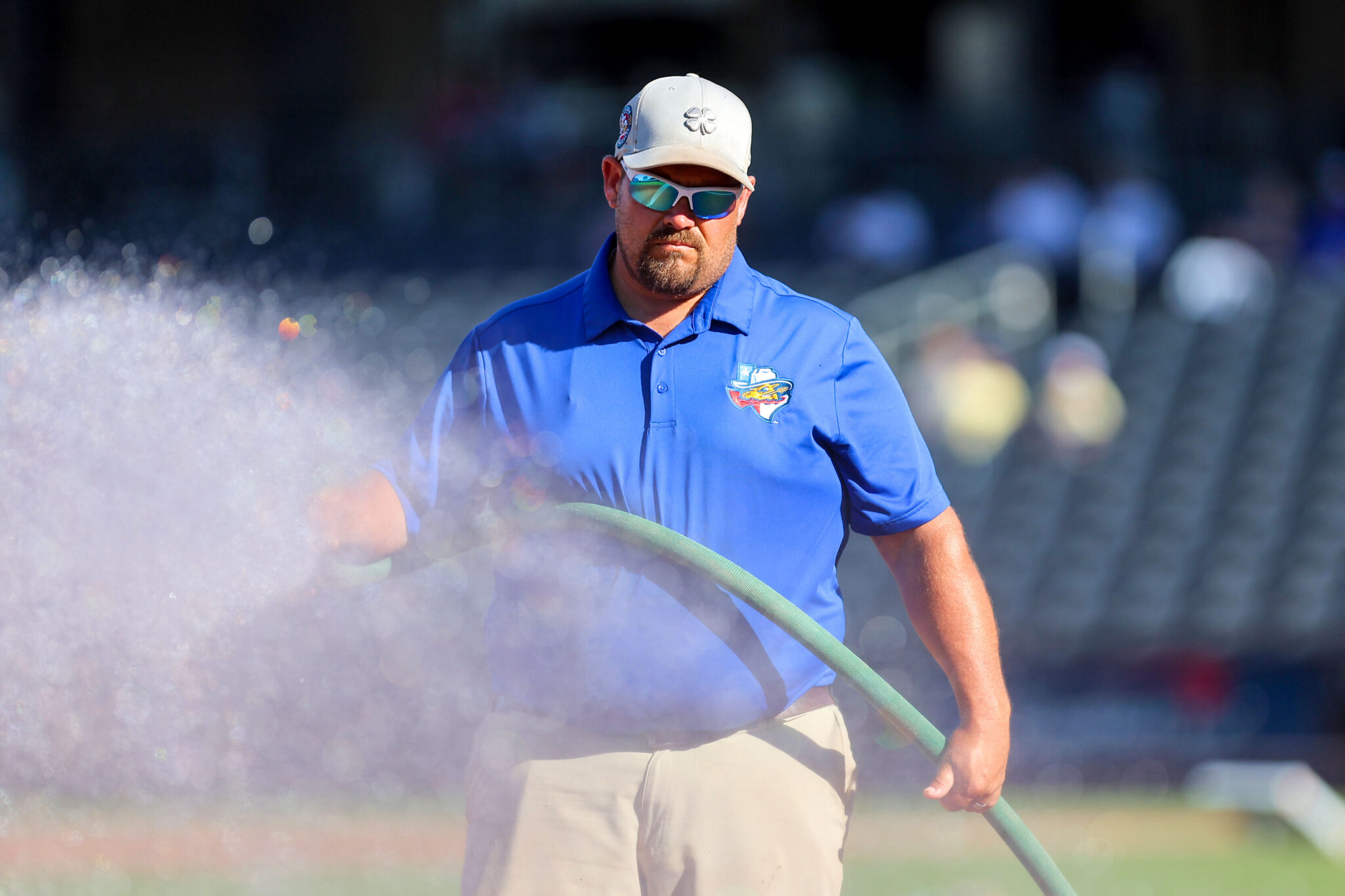 20260619-RockHounds-SodPoodles-0001 | Press Pass Sports Man in a blue polo shirt and beige cap with mirrored sunglasses, holding a green hose outdoors with misty air around him.