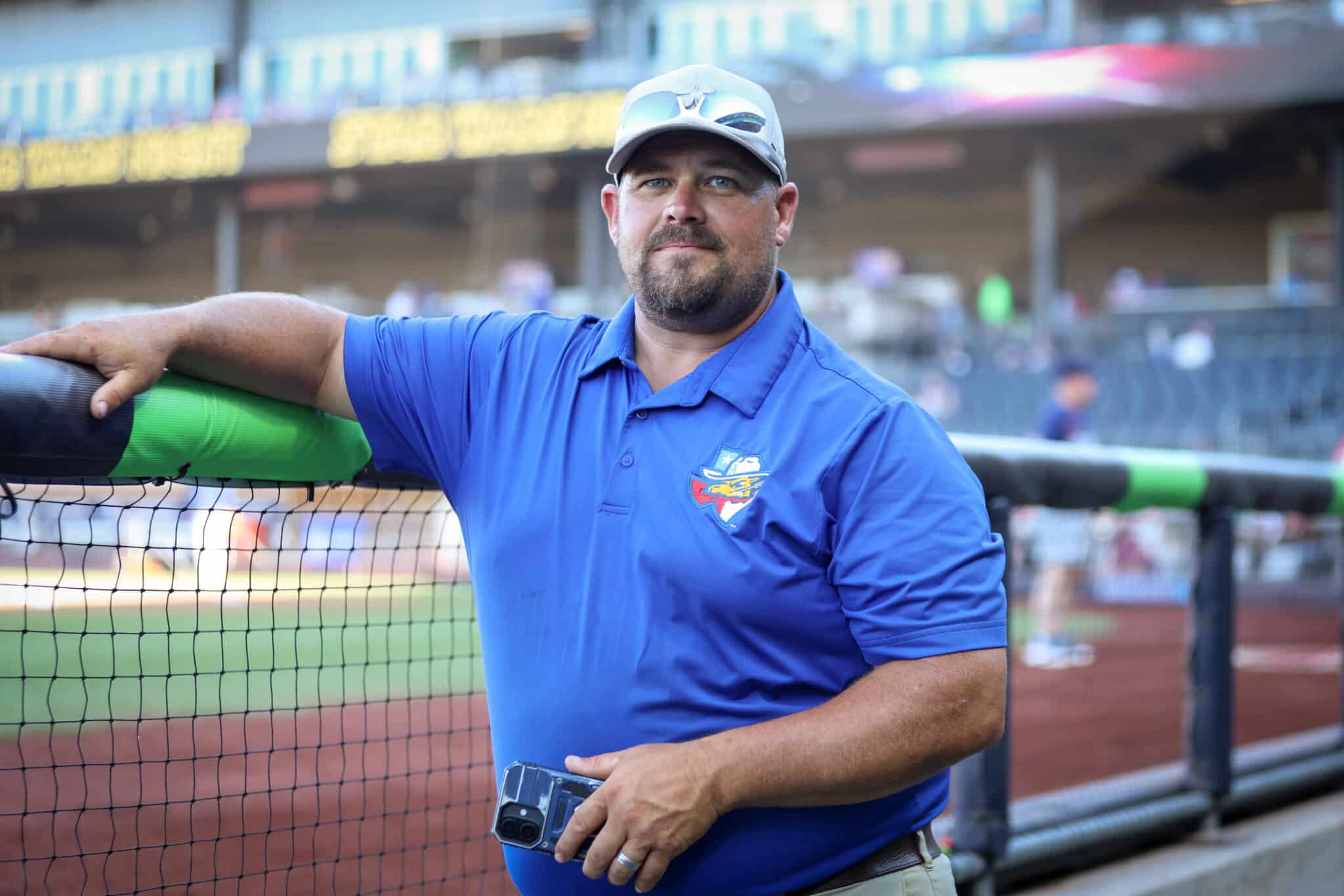 20260619-RockHounds-SodPoodles-0006 | Press Pass Sports Man in a blue polo shirt and cap rests his arm on a protective barrier at a baseball field behind a netting barrier.