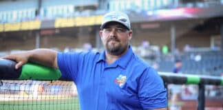 Man in a blue polo shirt and cap rests his arm on a protective barrier at a baseball field behind a netting barrier.