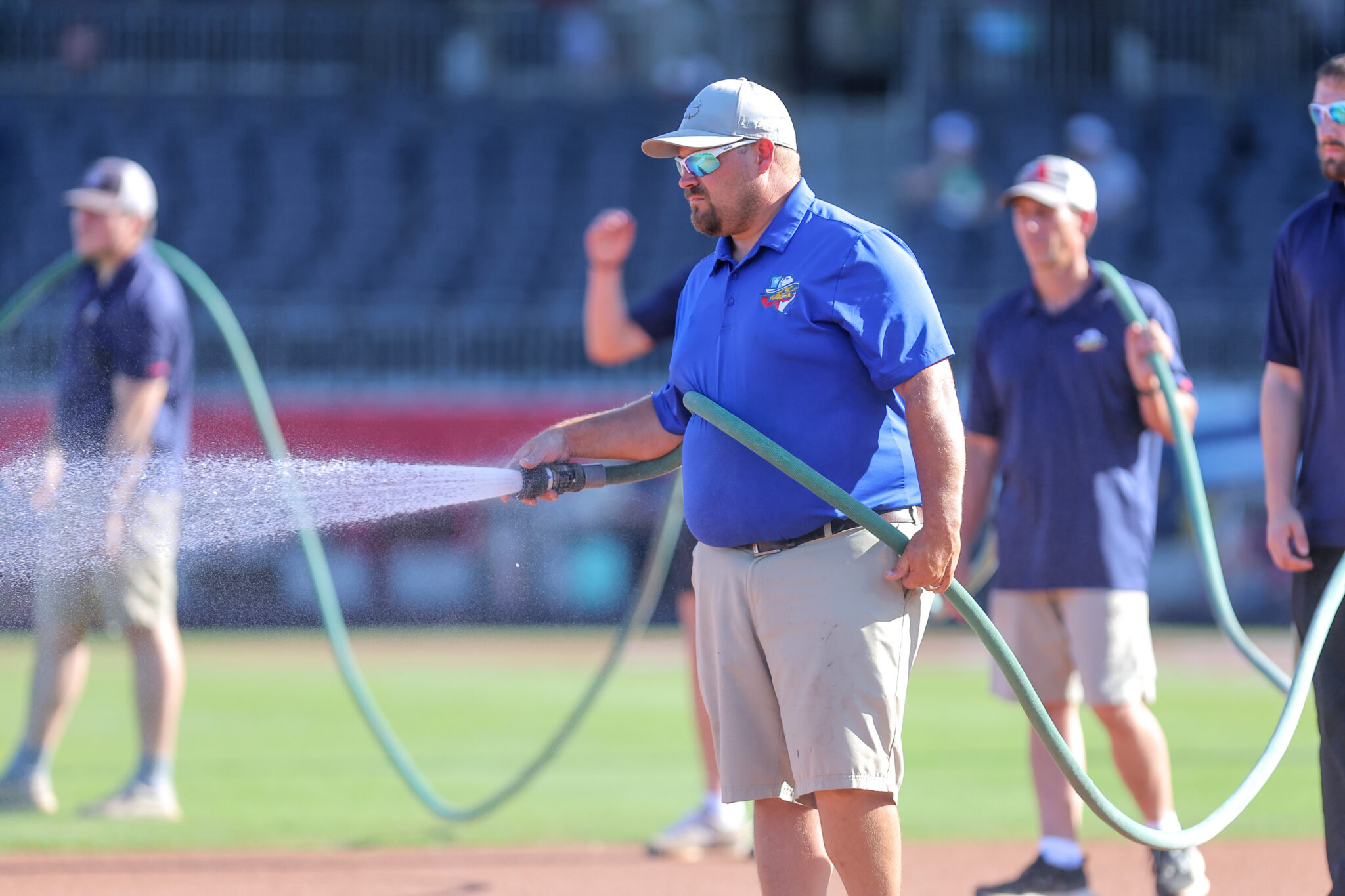 20260619-RockHounds-SodPoodles-0069 | Press Pass Sports Men in blue polo shirts watering a sports field with hoses during practice.