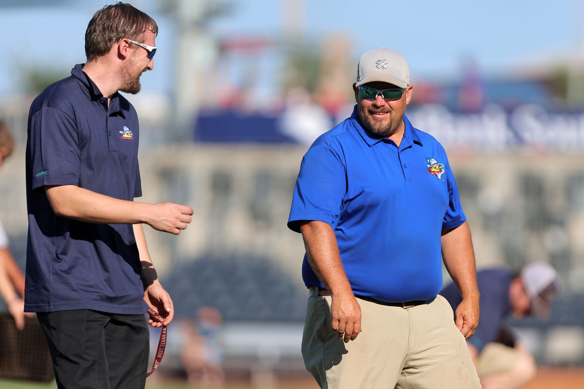 20260619-RockHounds-SodPoodles-0002 | Press Pass Sports Two men in polo shirts chat on a sunny sports field; one wears a navy shirt and sunglasses, the other a blue shirt and beige cap with a logo on it.
