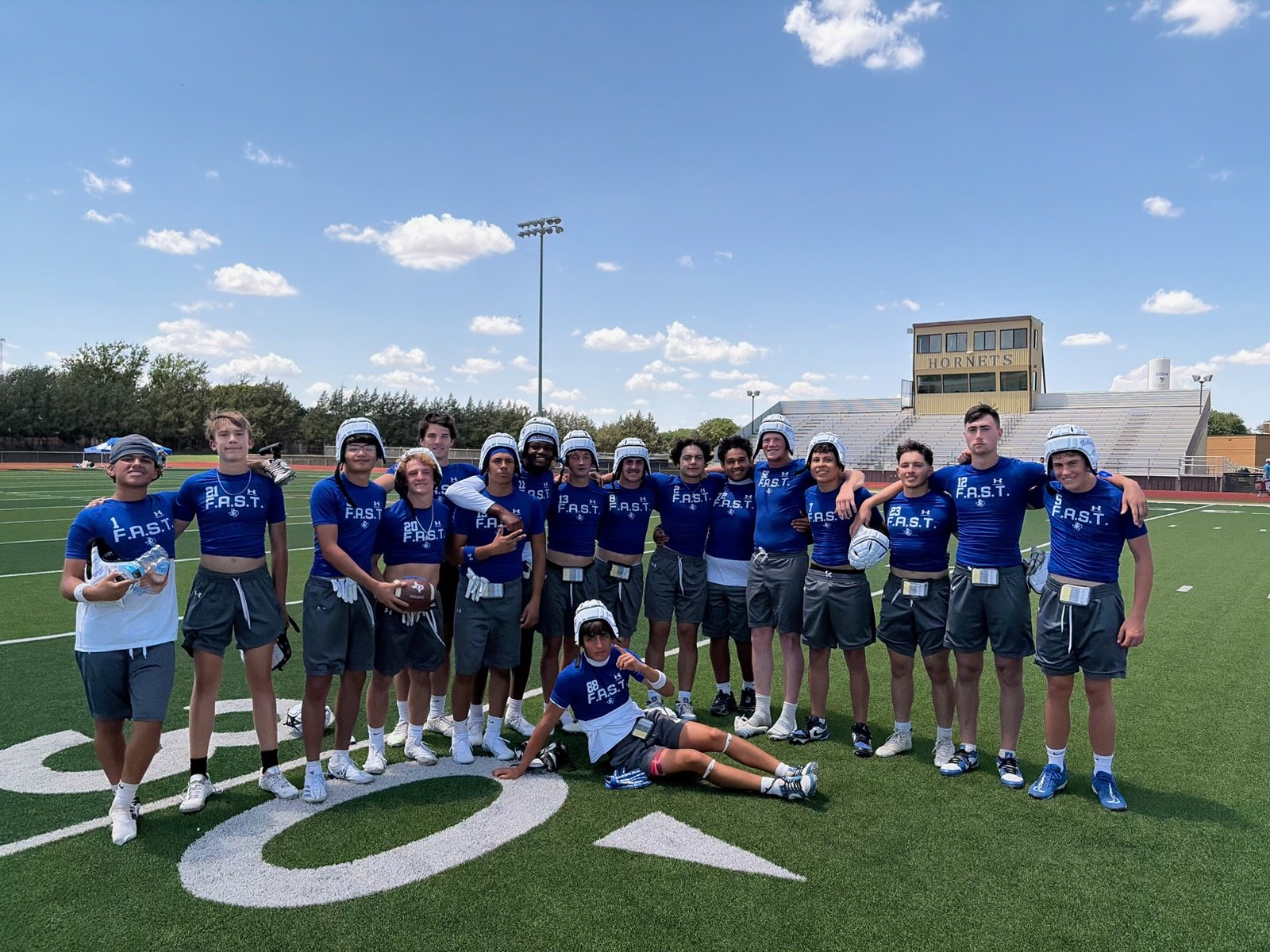 A group of young football players in blue FAST shirts and helmets posing together on a green field, with empty bleachers and a'HORNETS' building in the background.