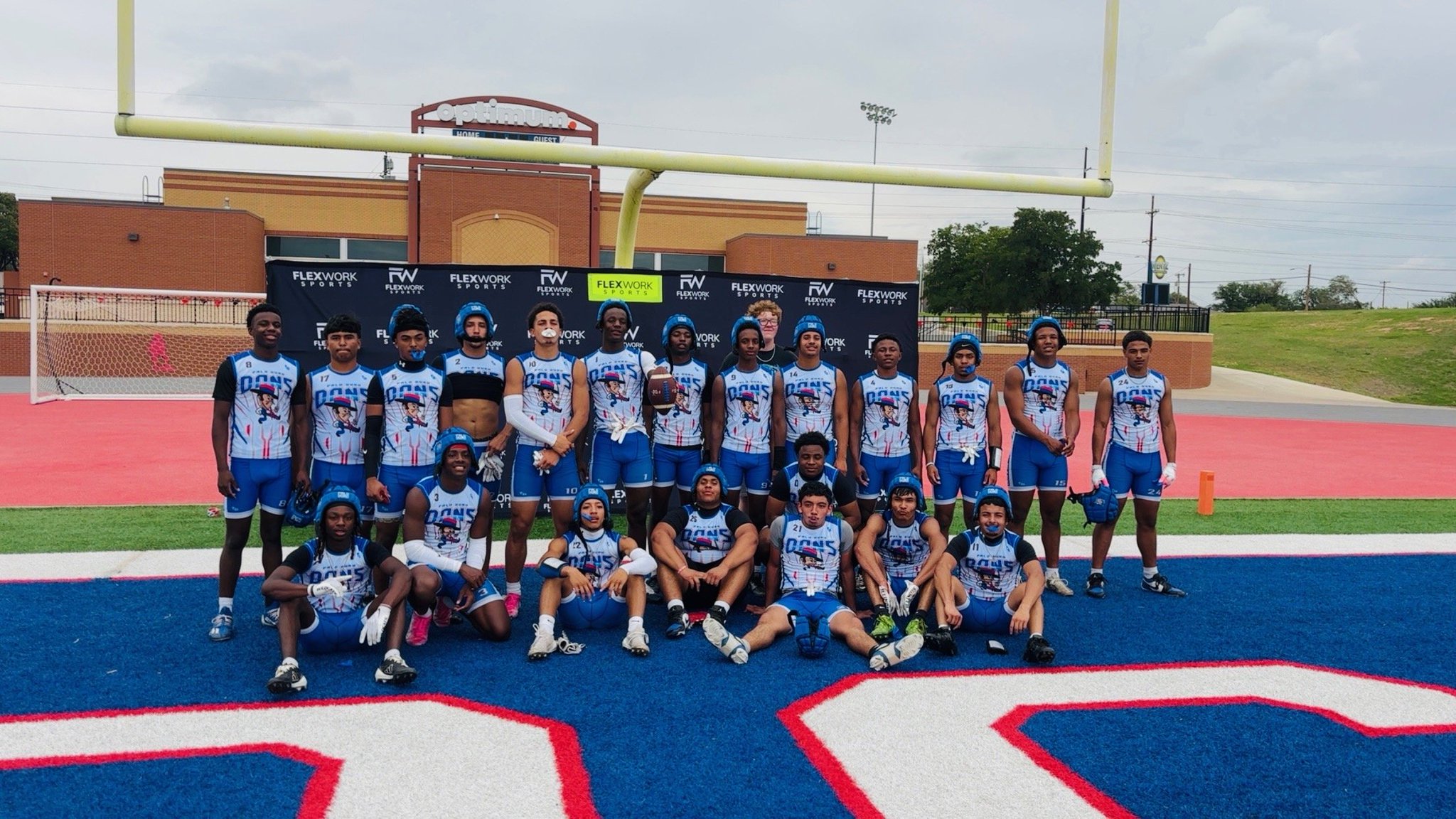 Group of youth football players in blue-and-white uniforms posing for a team photo on a blue turf field with a goalpost and stadium in the background.