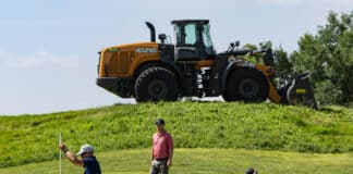 Golfers on a green putting while a large orange excavator sits on a hill behind them.