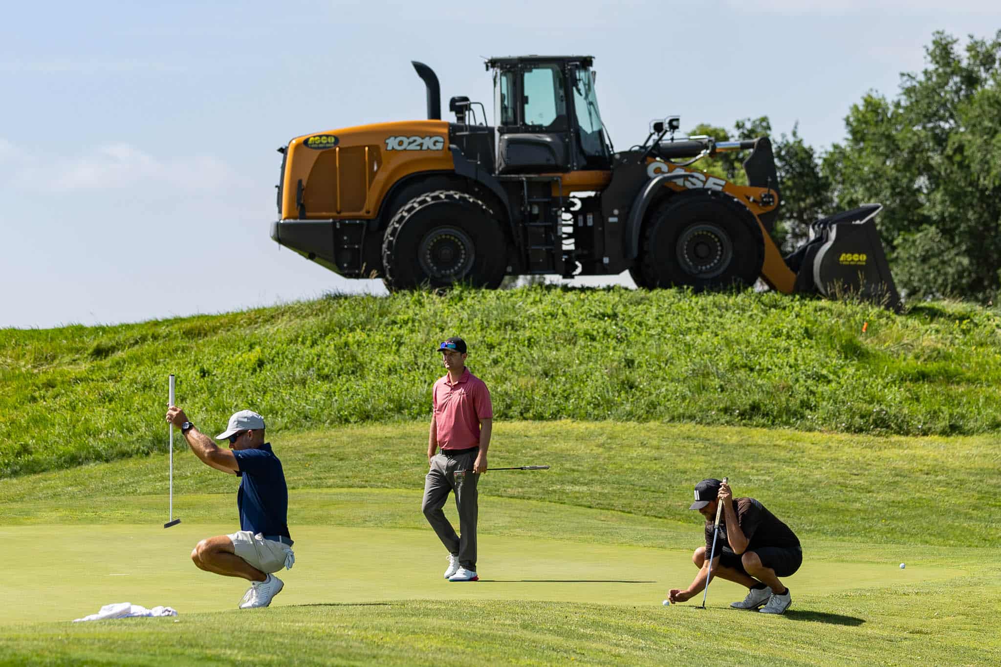 Golfers on a green putting while a large orange excavator sits on a hill behind them.