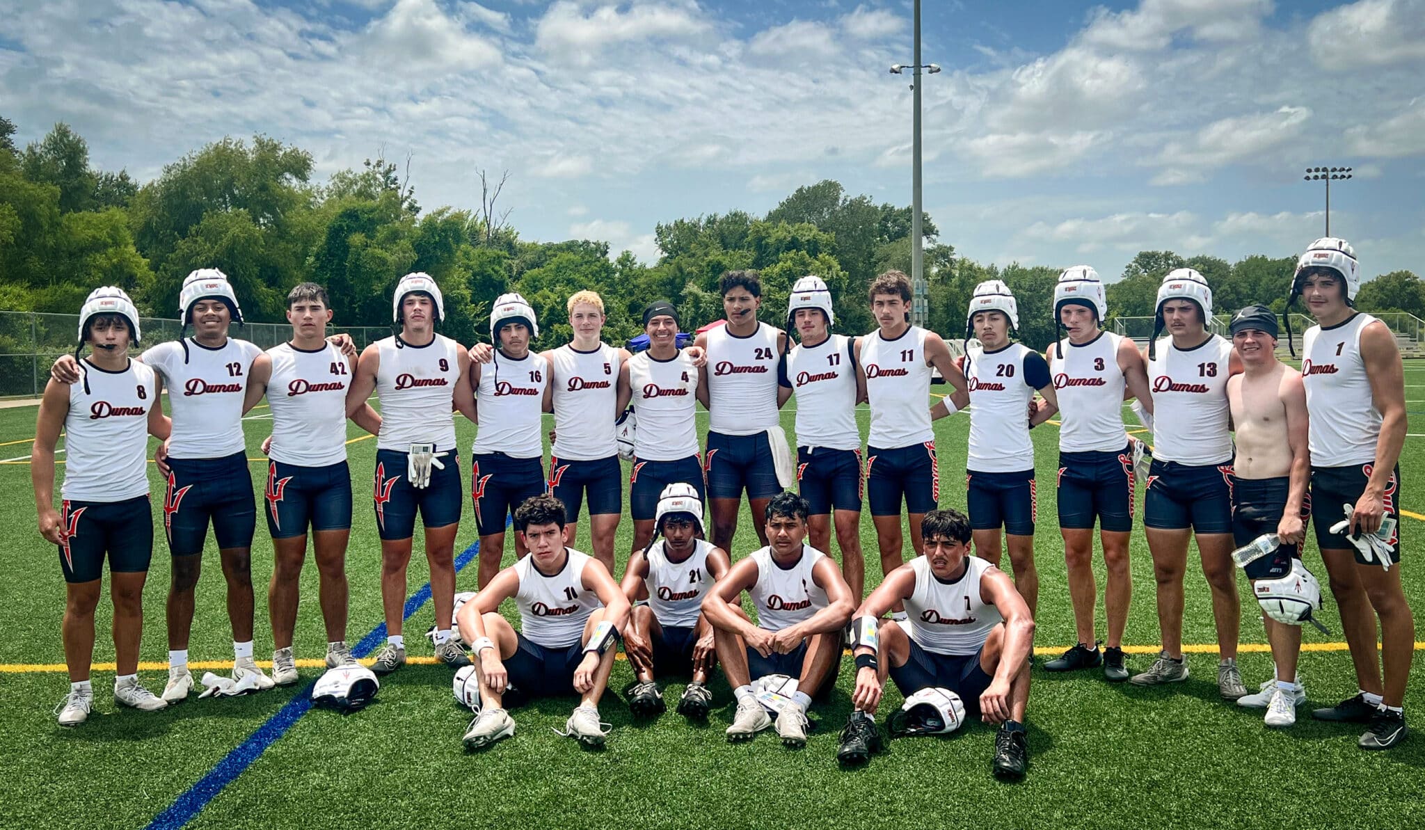 Group of male lacrosse players in white Dumas jerseys posing together on a field, wearing helmets and shorts.