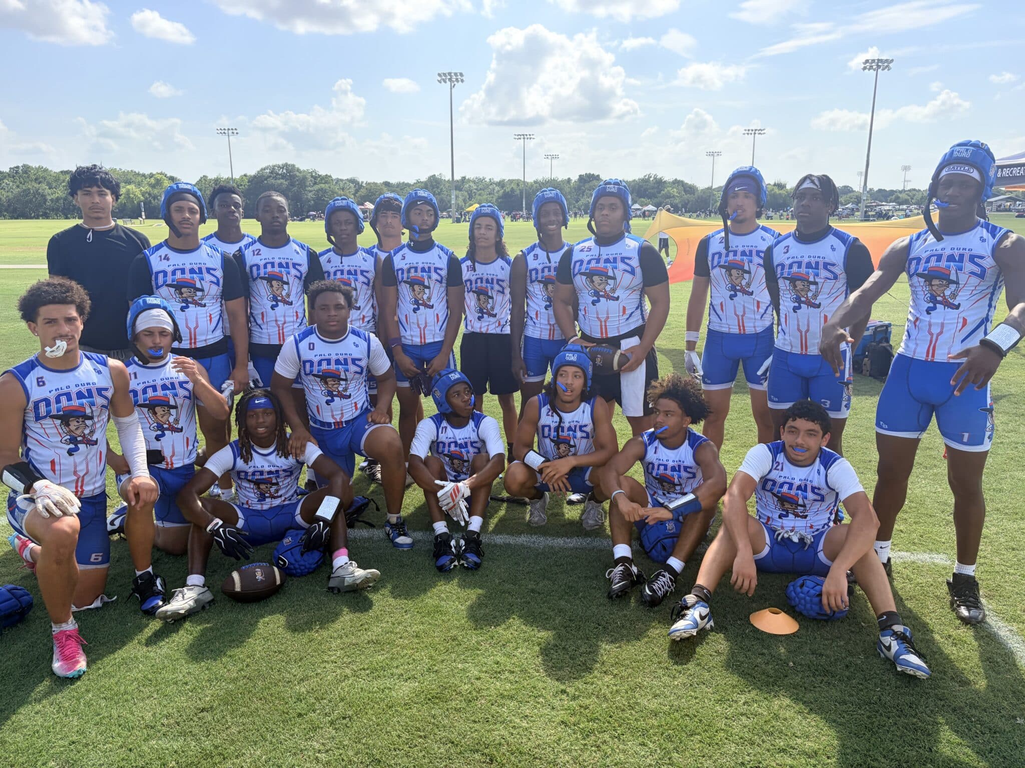 Group of young football players in blue-and-white uniforms posing on a sunny practice field with a coach nearby.