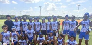 Group of young football players in blue-and-white uniforms posing on a sunny practice field with a coach nearby.