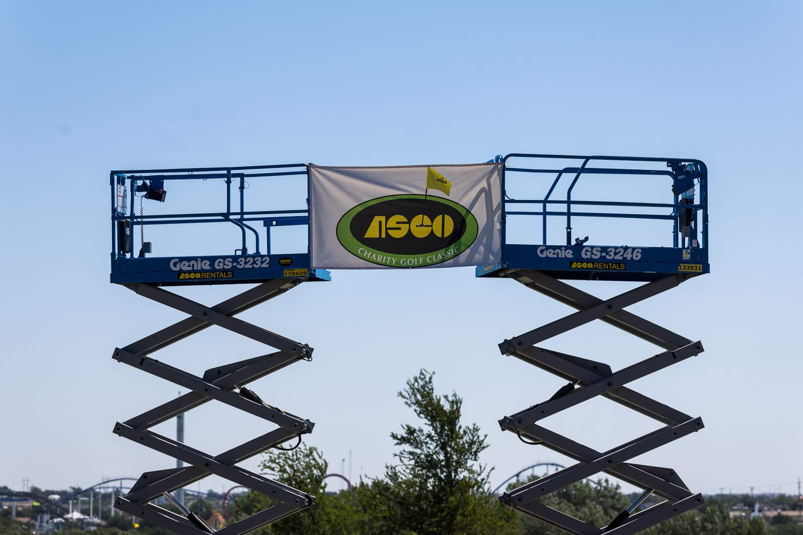 Two blue scissor lifts raised high form a bridge holding a banner for the ASCO Charity Golf Classic.
