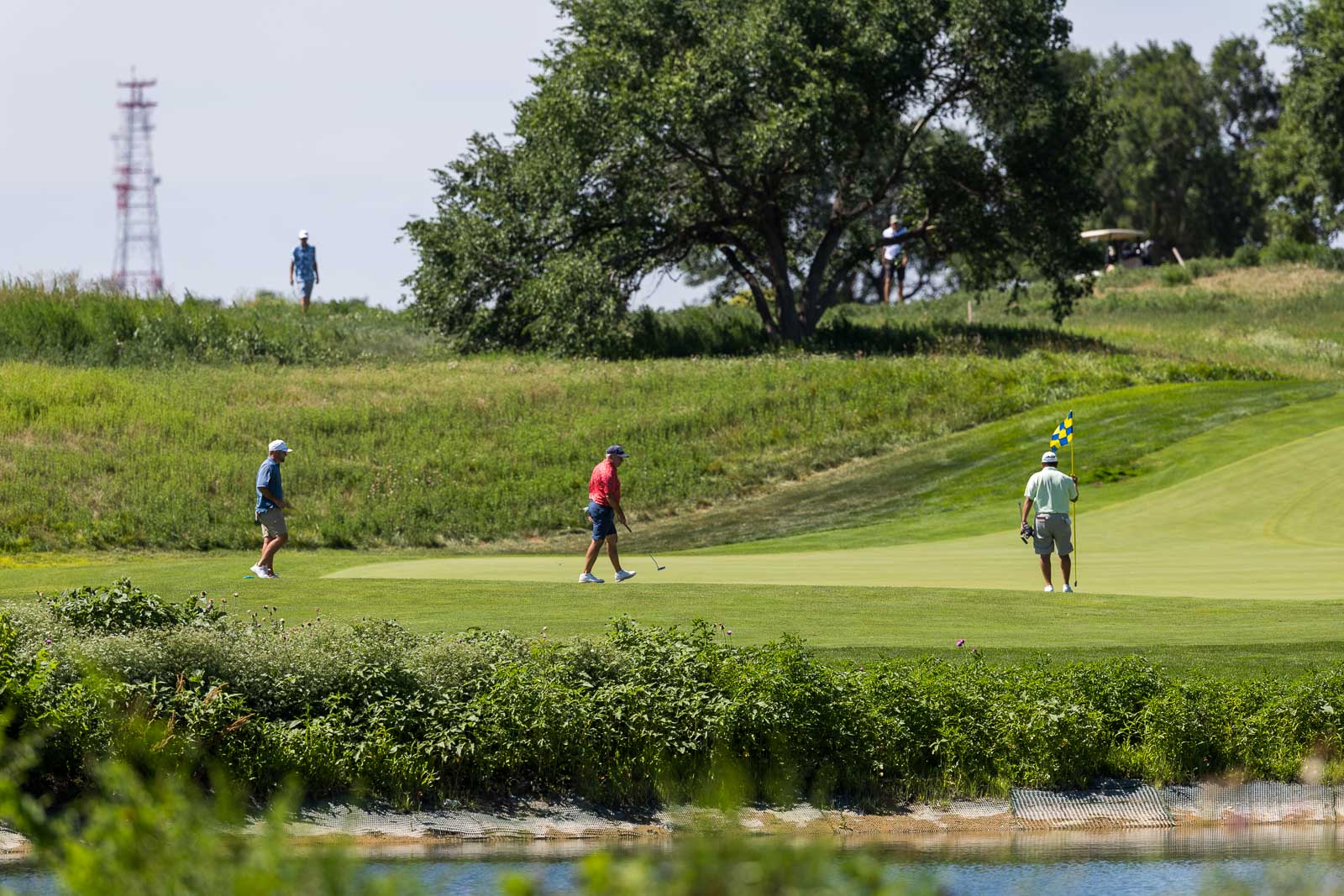Golf course putting green with three players; one man in red prepares a putt while another holds the flag near the hole.