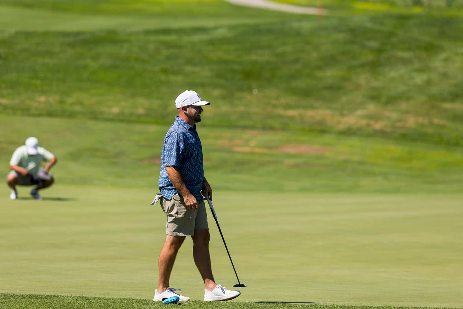 Golfer in a blue patterned shirt and white cap walking on the green with a putter while a companion kneels in the background.