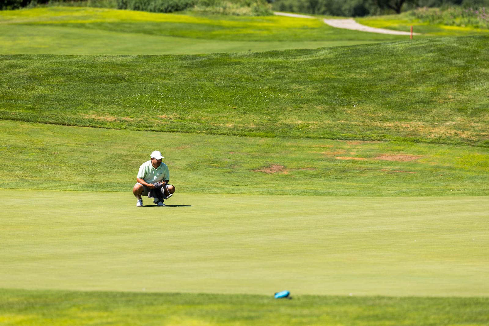 Golfer crouching on a putting green, lining up a shot with a club in hand under bright sun