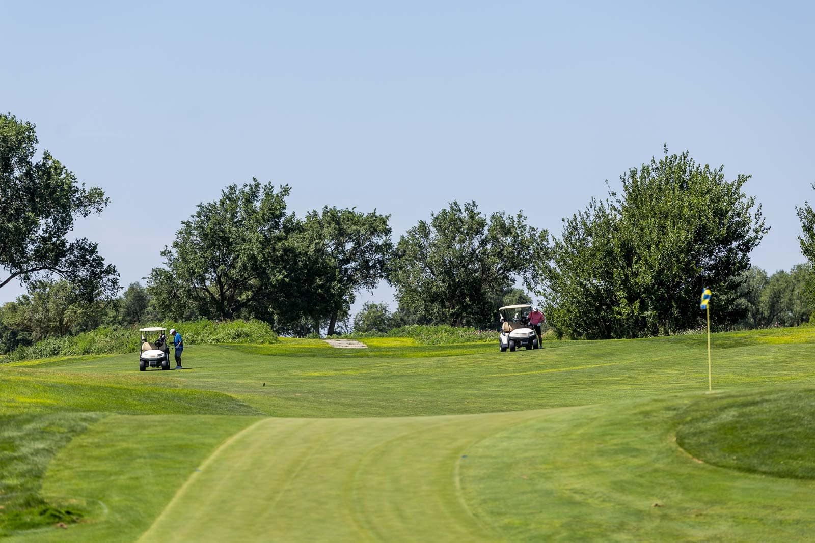 Two golfers on a sunny golf course near a cart path, with two golf carts and a green flag in the distance.