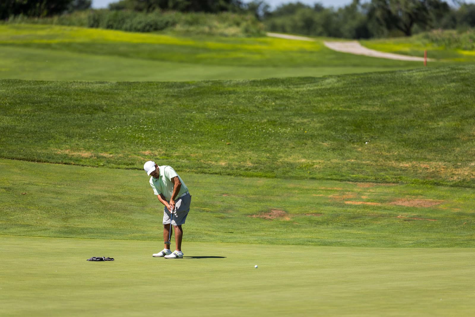 Golfer in a white cap and light-green shirt lines up a putt on a green, with a ball nearby and a grassy landscape behind.