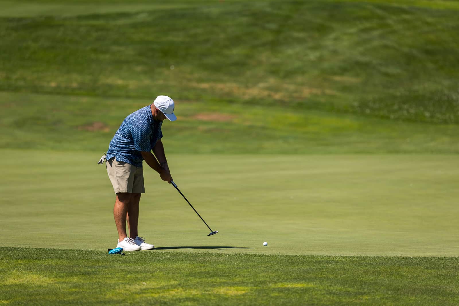 Man in a blue patterned shirt, khaki shorts, and white cap lining up a putt on a golf green.