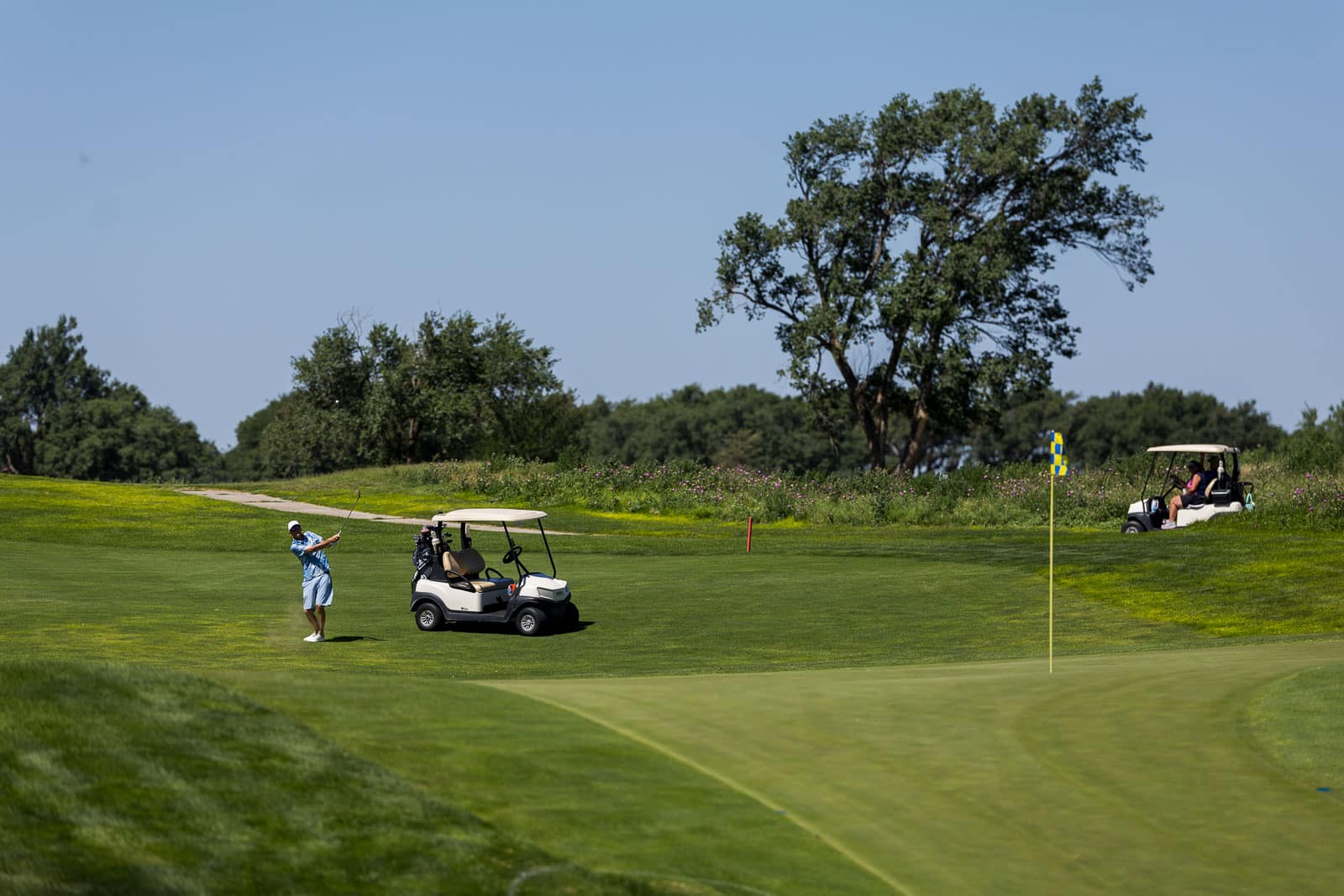 Golfer in a blue shirt and white cap mid-swing on a sunny golf course, with a golf cart nearby.