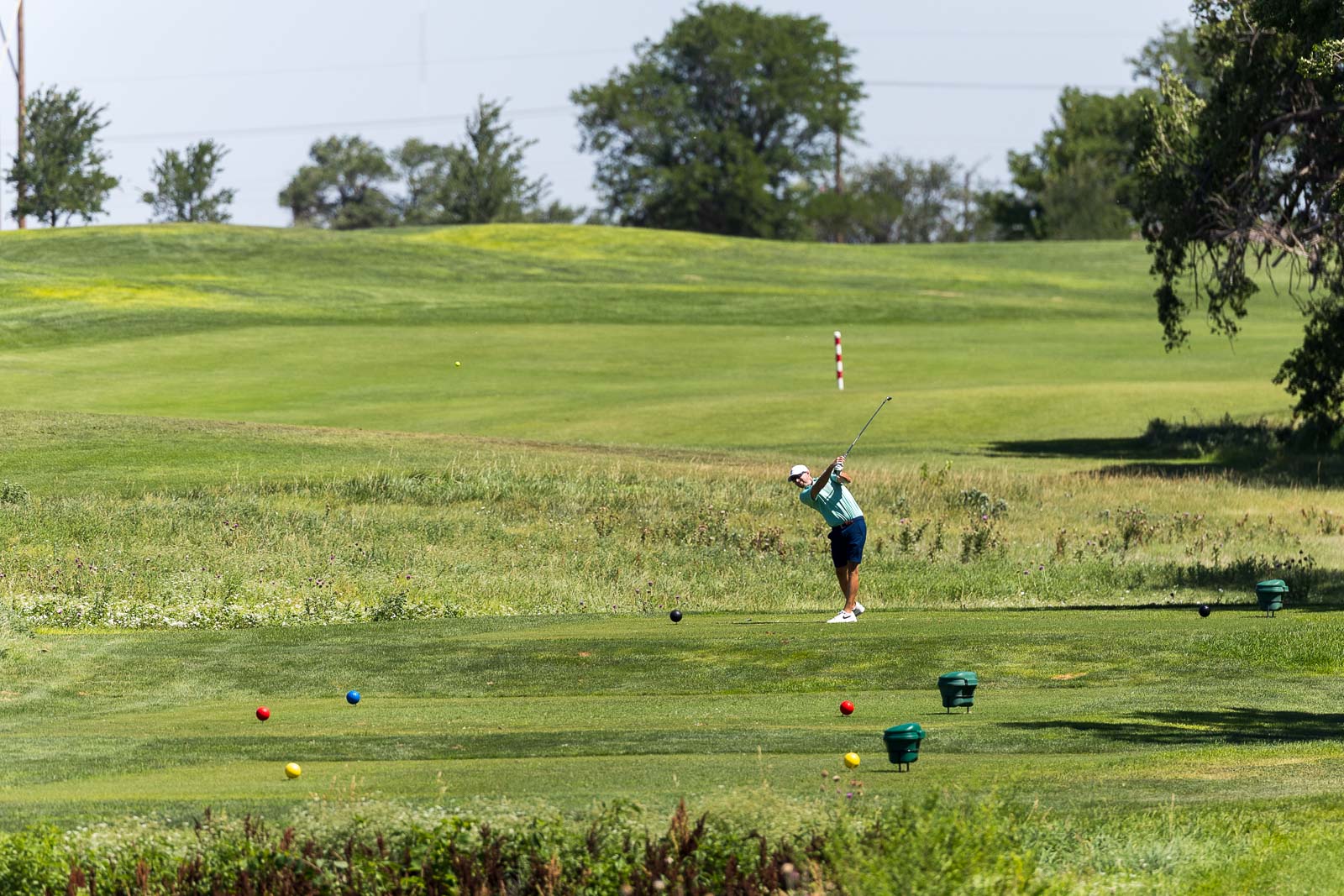 Golfer in a teal shirt and white cap mid-swing on a sunny driving range, with colorful practice balls on the grass in front of him.