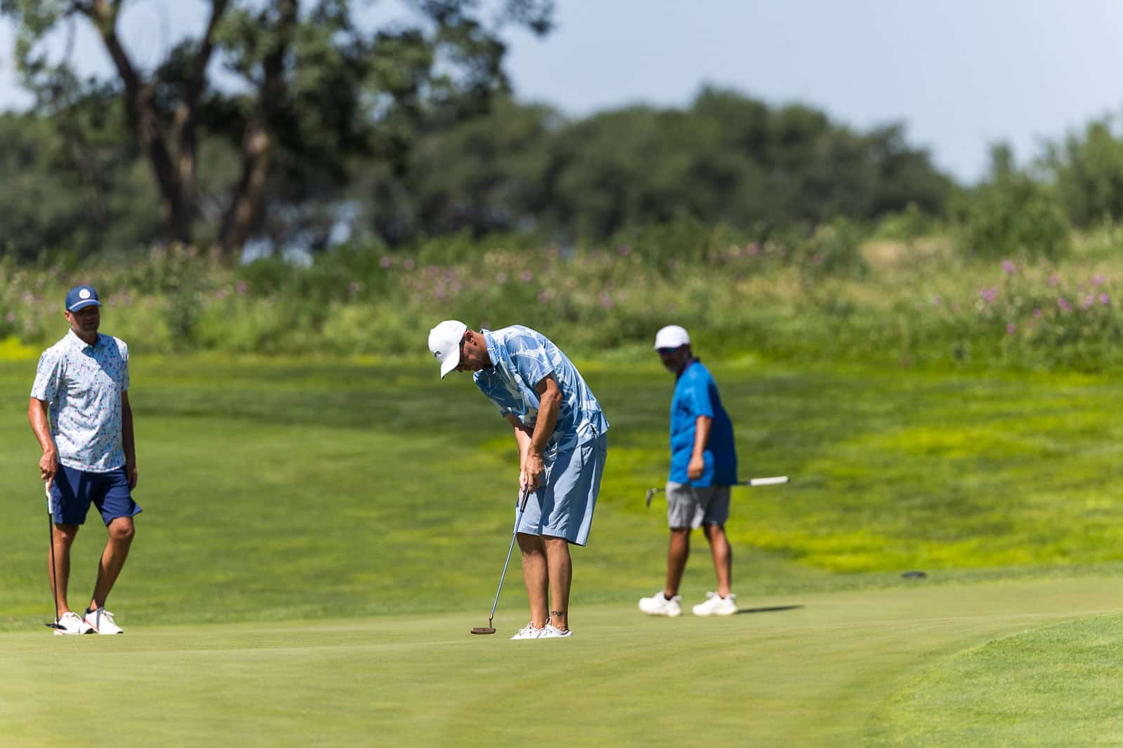 Golfer in a blue patterned shirt puts on the green while two teammates watch in the background on a sunny course.