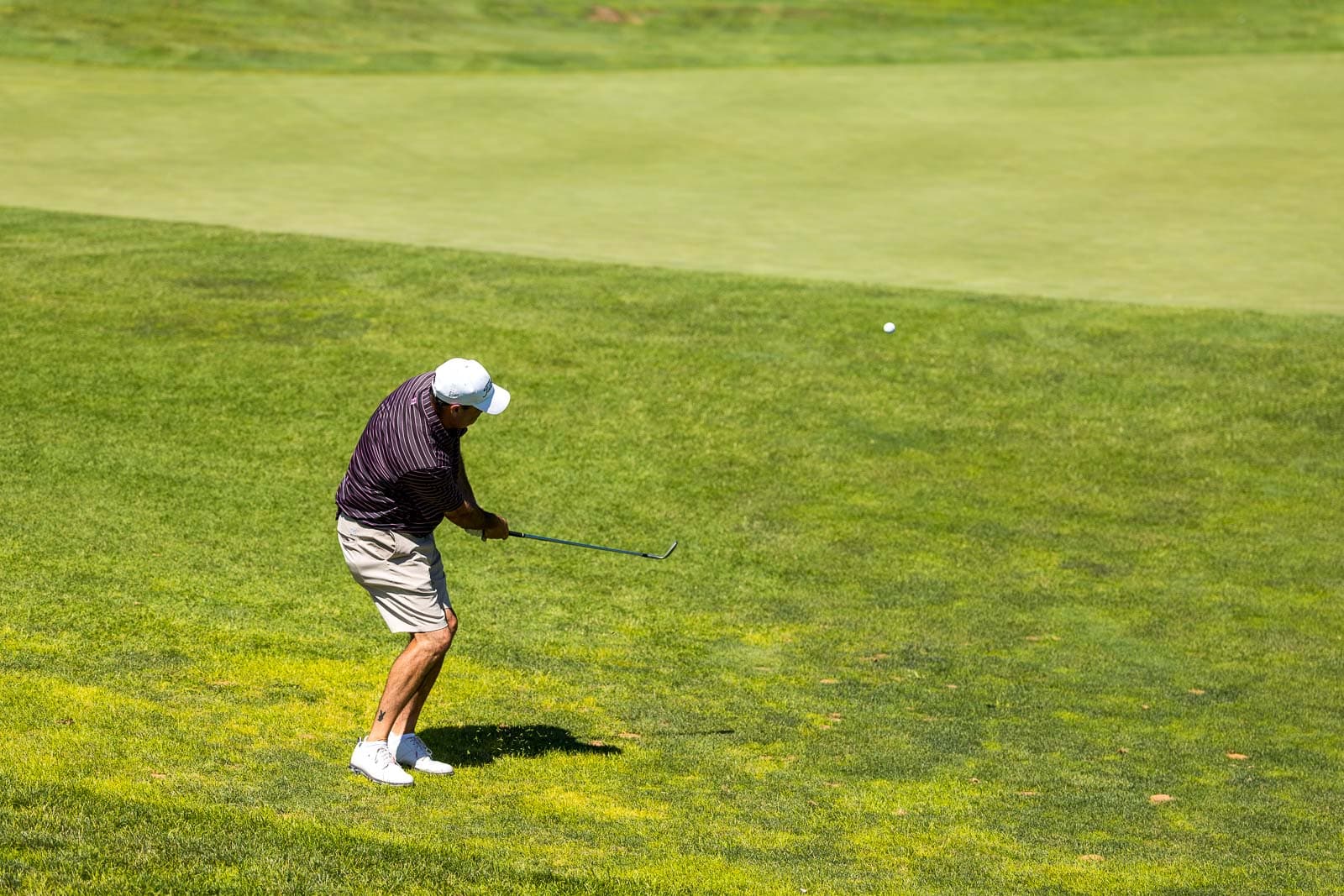 Golfer in a striped shirt and white cap swings a club on a green golf course, ball in midair.