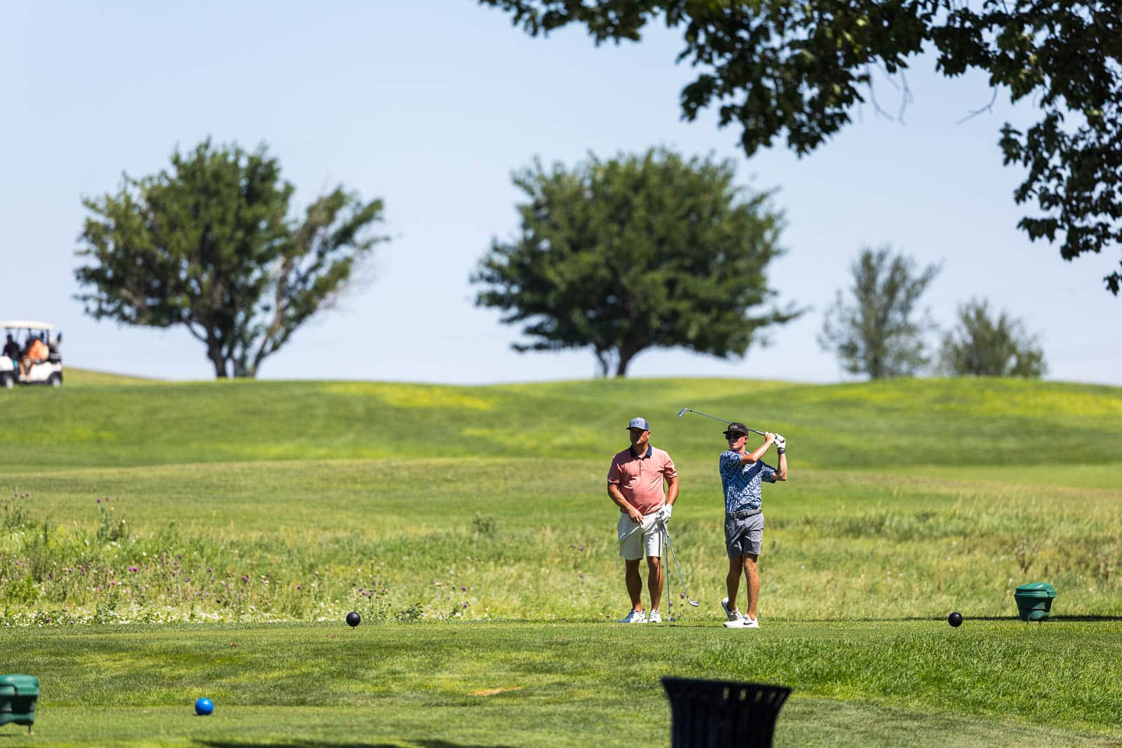 Two golfers on a sunny course, one swinging a club while the other watches nearby.