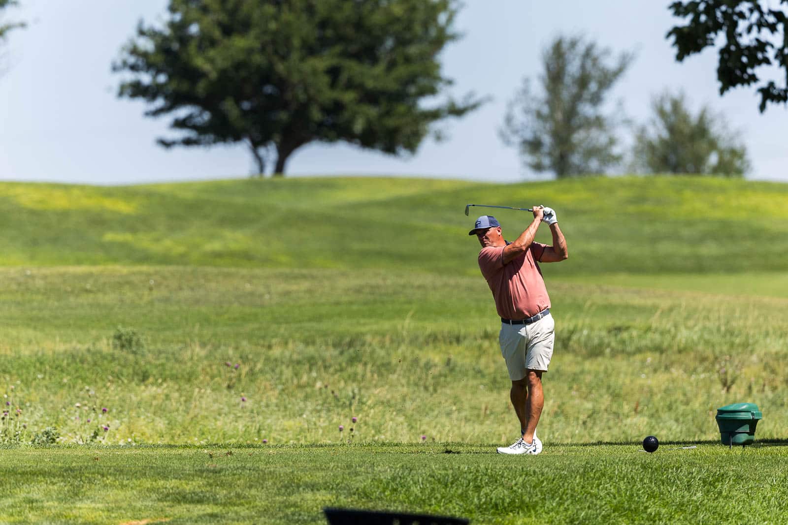 Golfer in a pink shirt and cap mid-swing on a sunny golf course with green fairways and trees in the background.