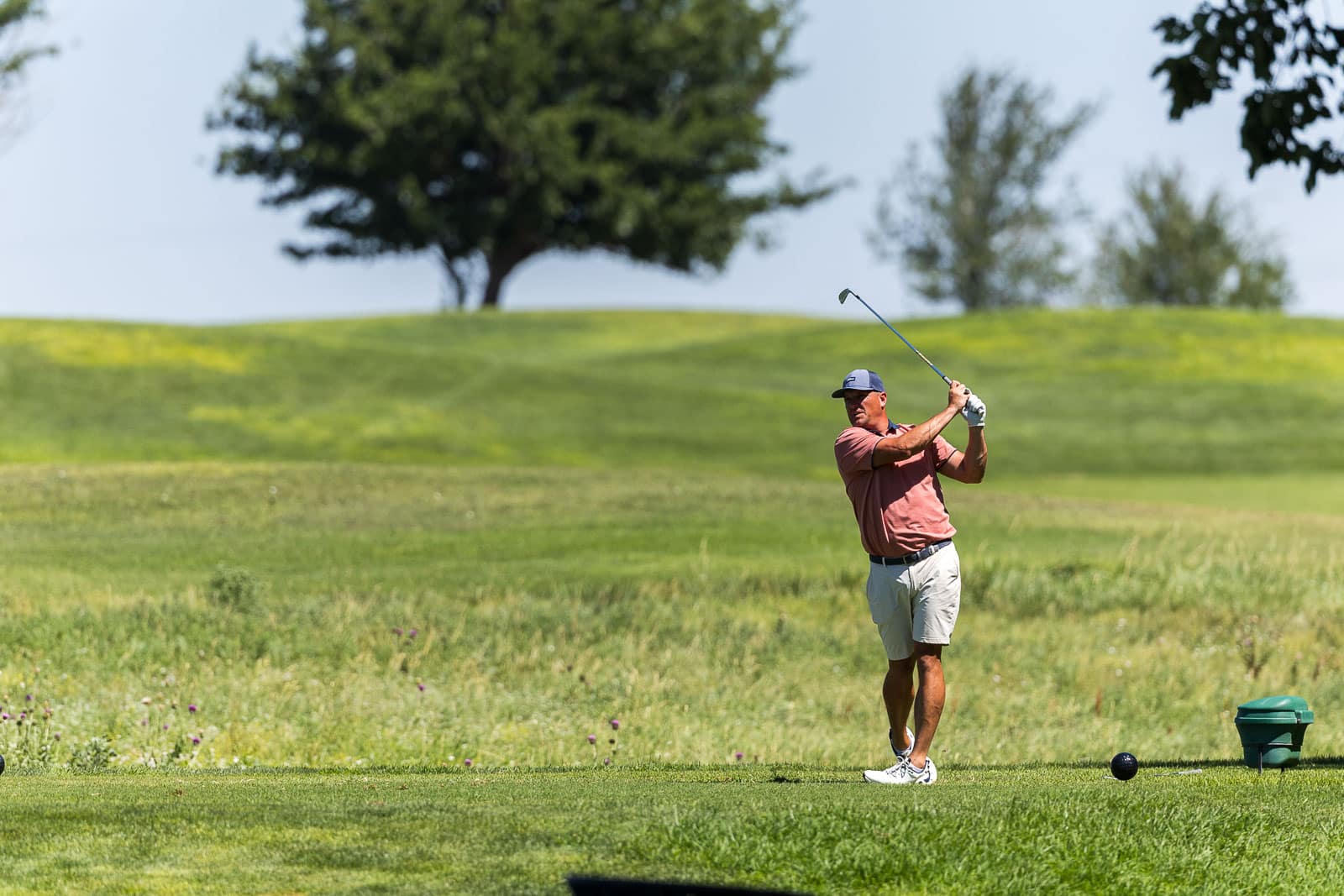 Golfer mid-swing on a sunny green golf course with trees in the background, wearing a pink shirt and cap.