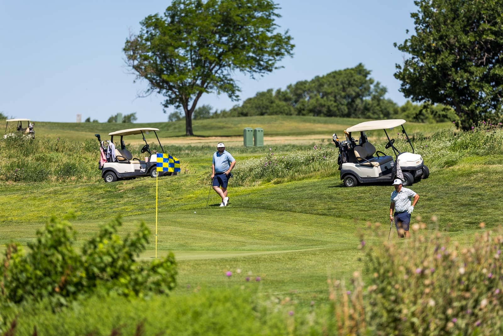 Two golfers on a sunny golf course near a flagstick with a cresting green and rolling fairways, two golf carts parked nearby.
