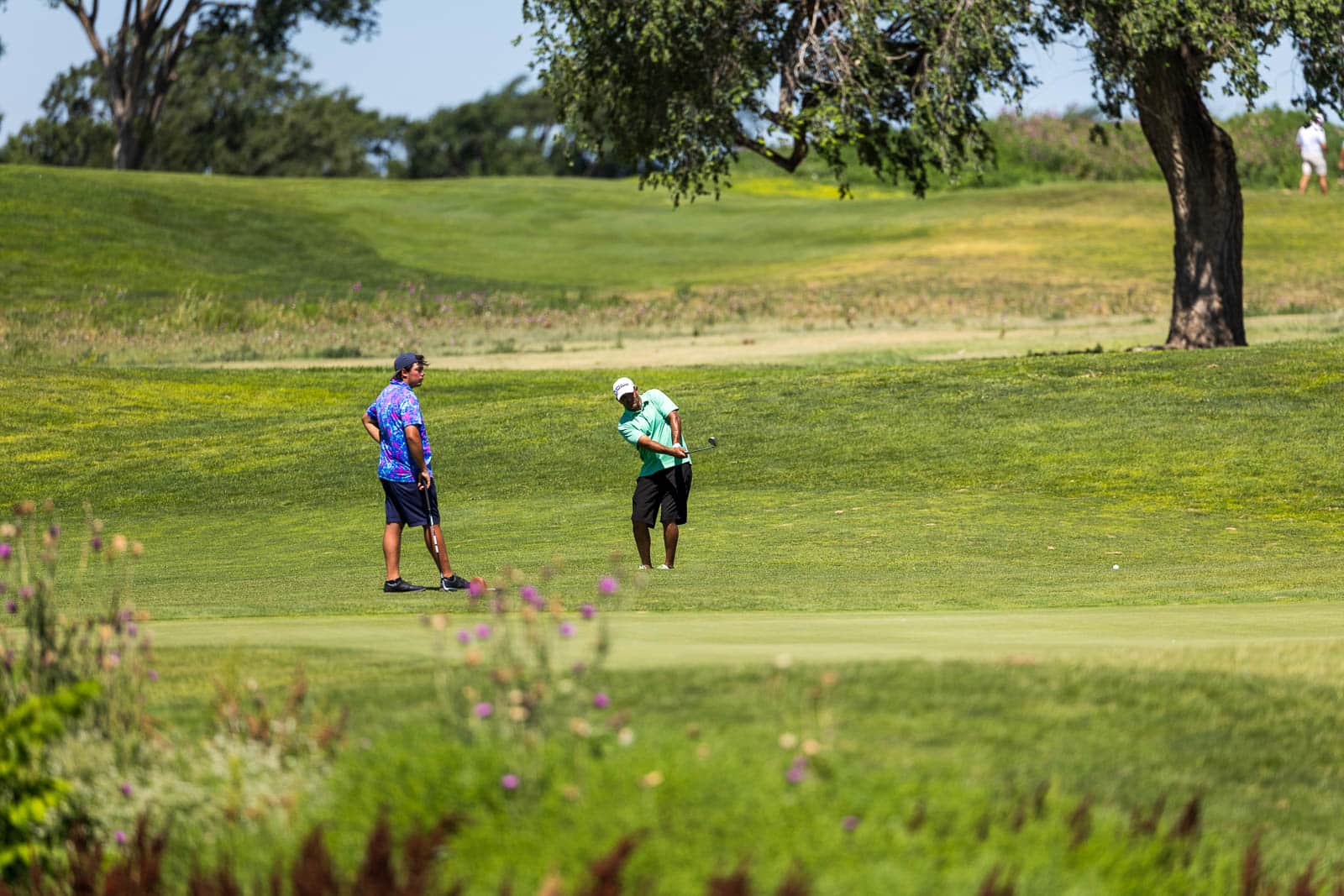 Two golfers on a sunny golf course: one in a mint-green shirt mid-swing, the other in a bright patterned shirt watches nearby.